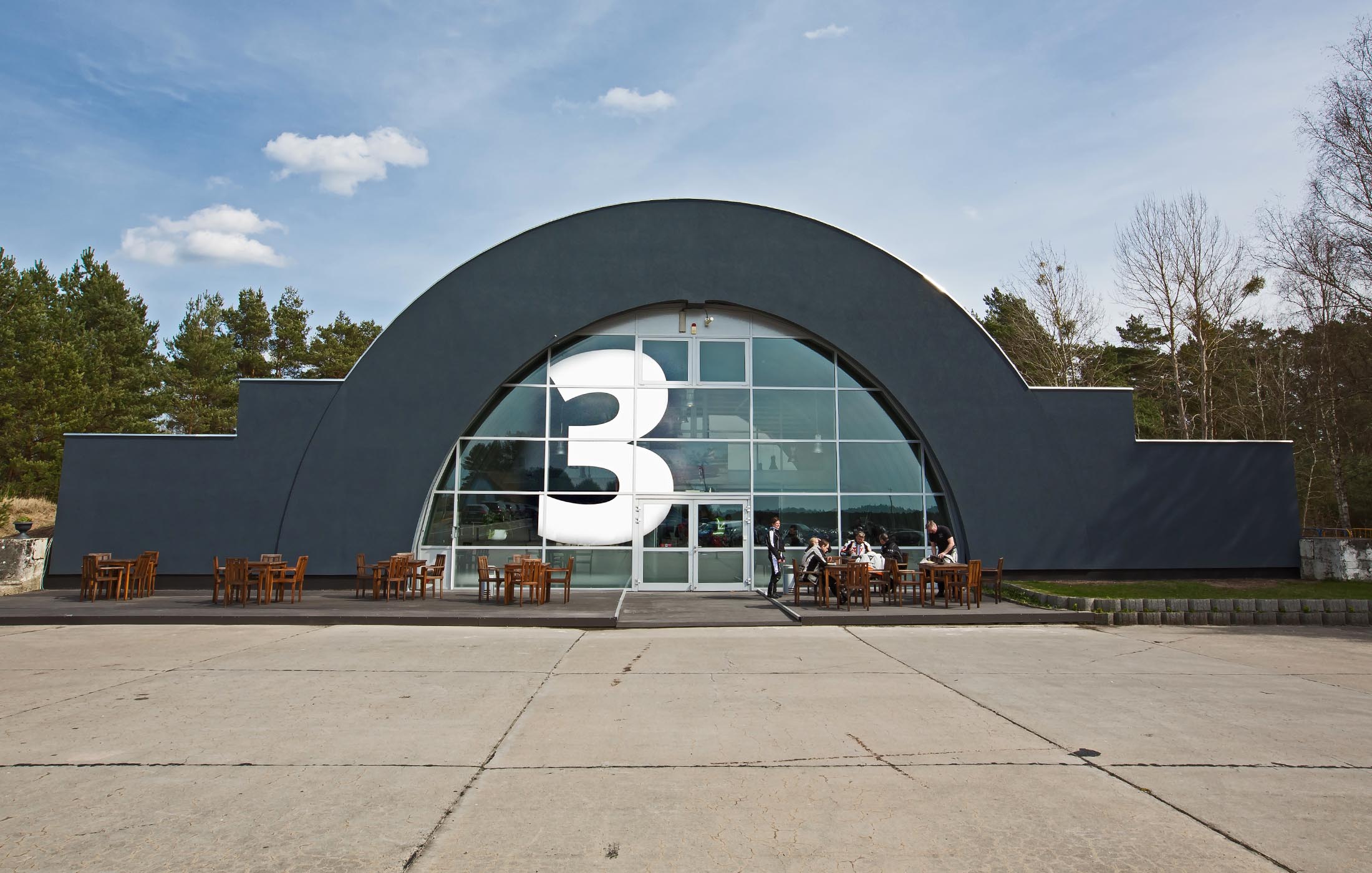 Frontansicht eines modernen Gebäudes mit großer Nummer drei an der Glasfassade und Holzsitzgruppen davor.