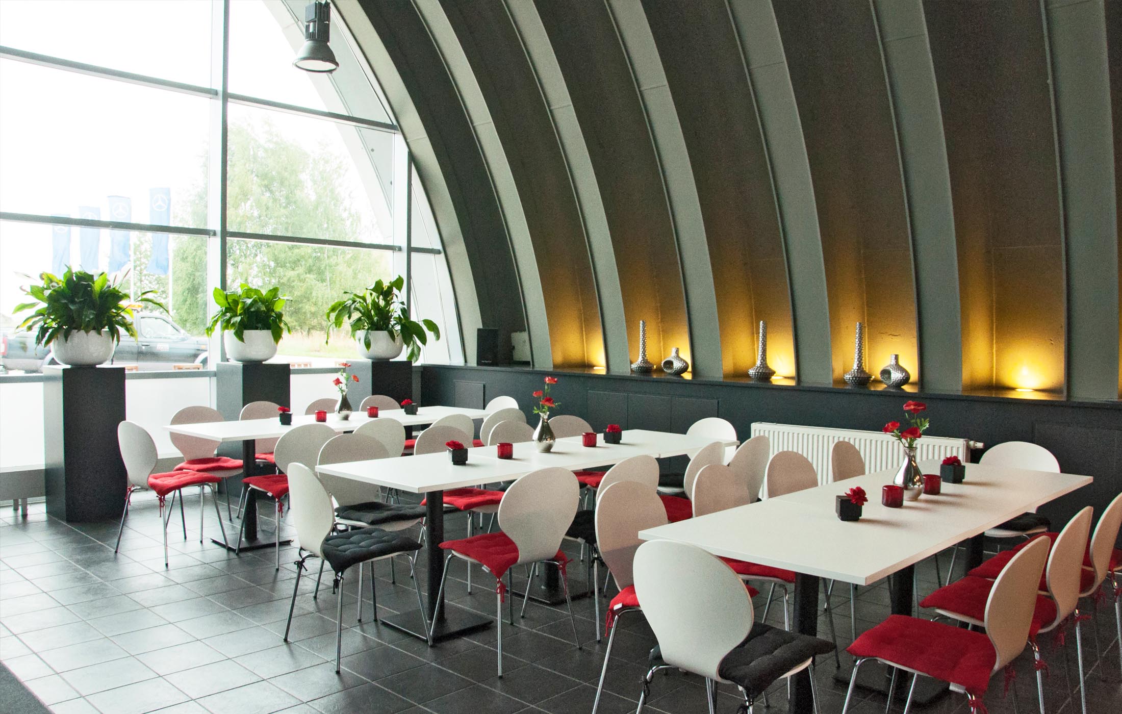 Blick von oben auf ein modernes, lichtdurchflutetes Bistro mit weißen Tischen, roten Sitzkissen und großen Pflanzen vor Panoramafenstern.
