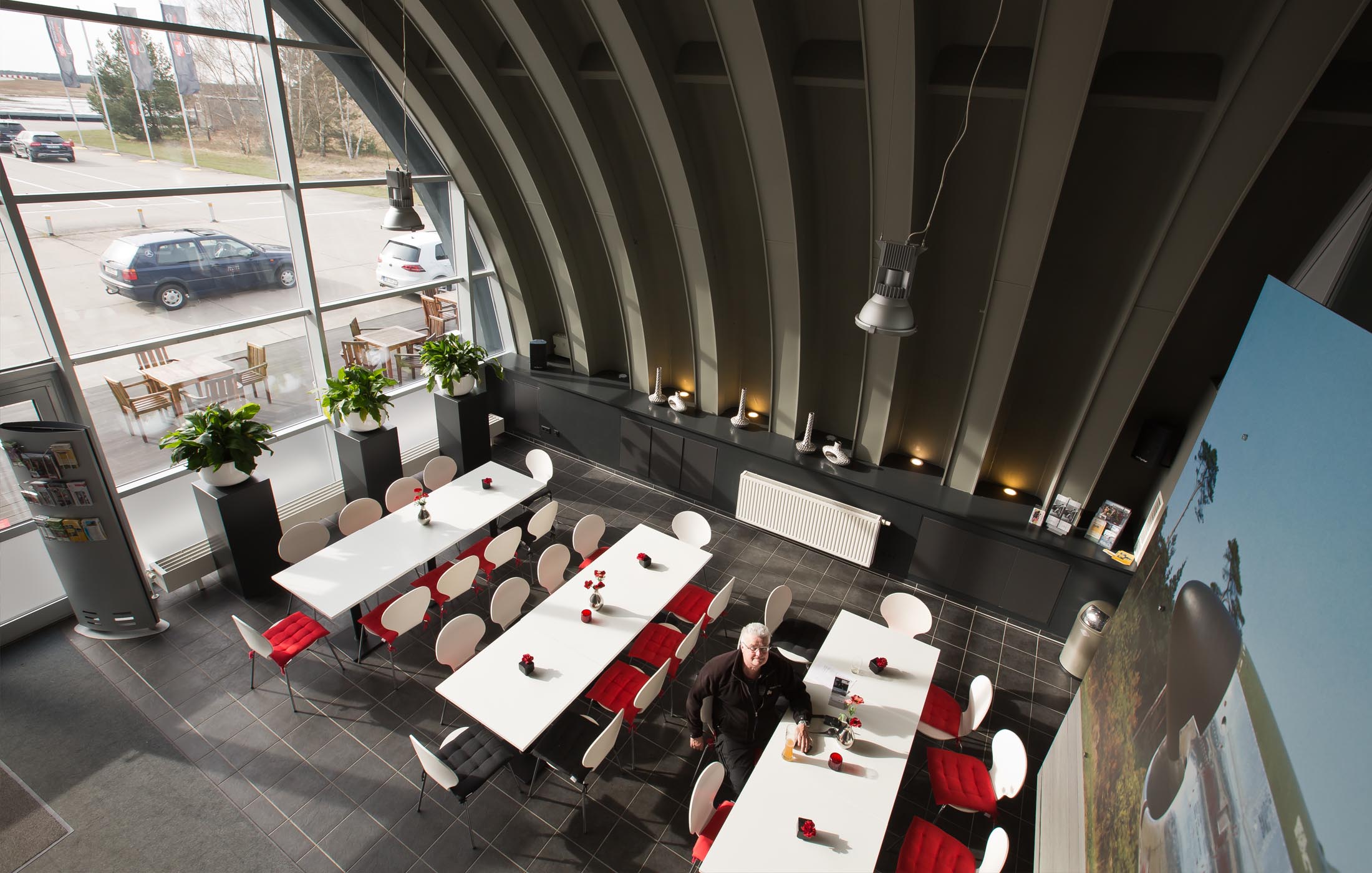 Blick von oben auf ein modernes, lichtdurchflutetes Bistro mit weißen Tischen, roten Sitzkissen und großen Pflanzen vor Panoramafenstern.