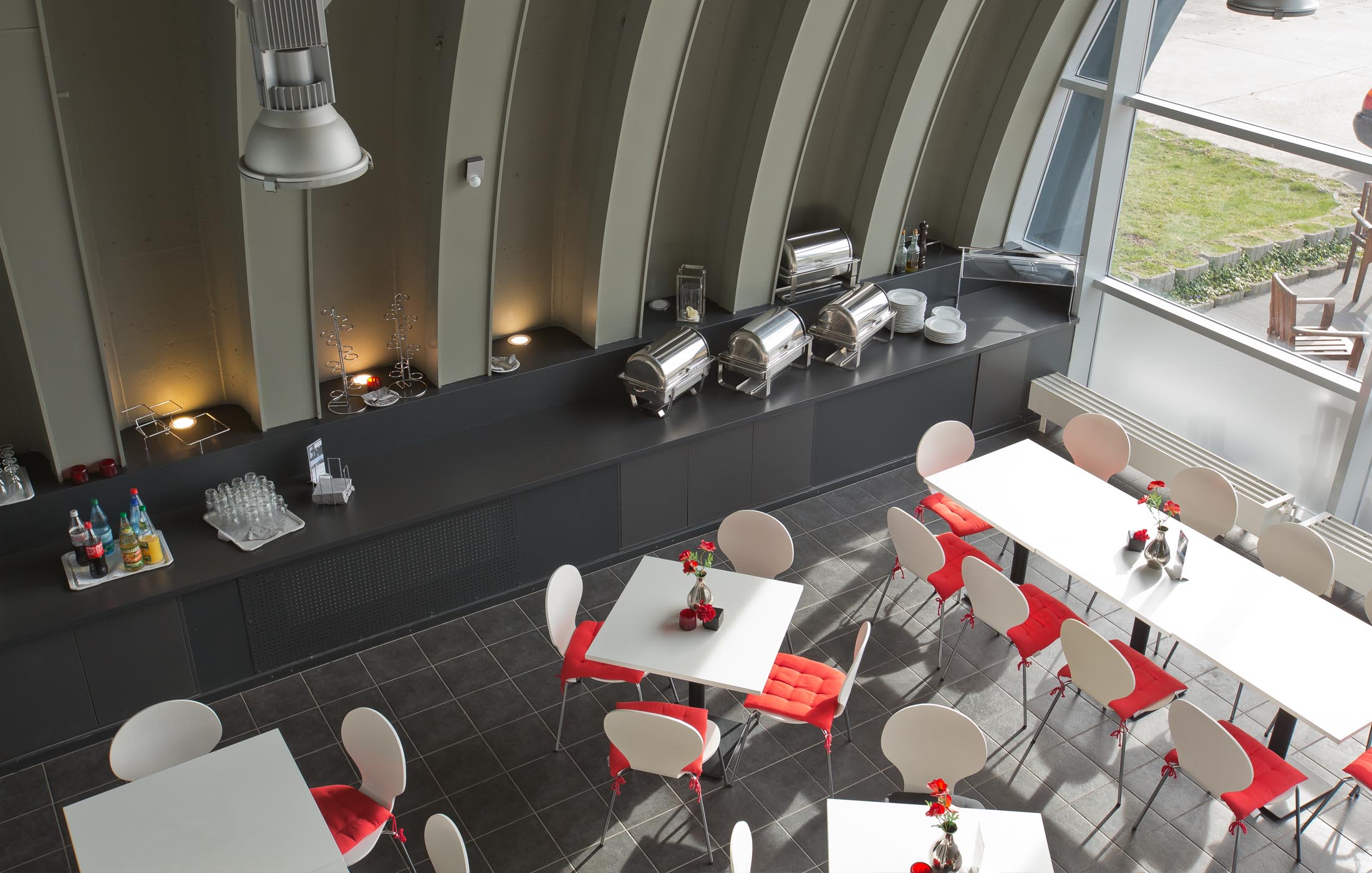 Moderner Kantinenbereich mit weißen Tischen, roten Sitzkissen und großer Fensterfront.