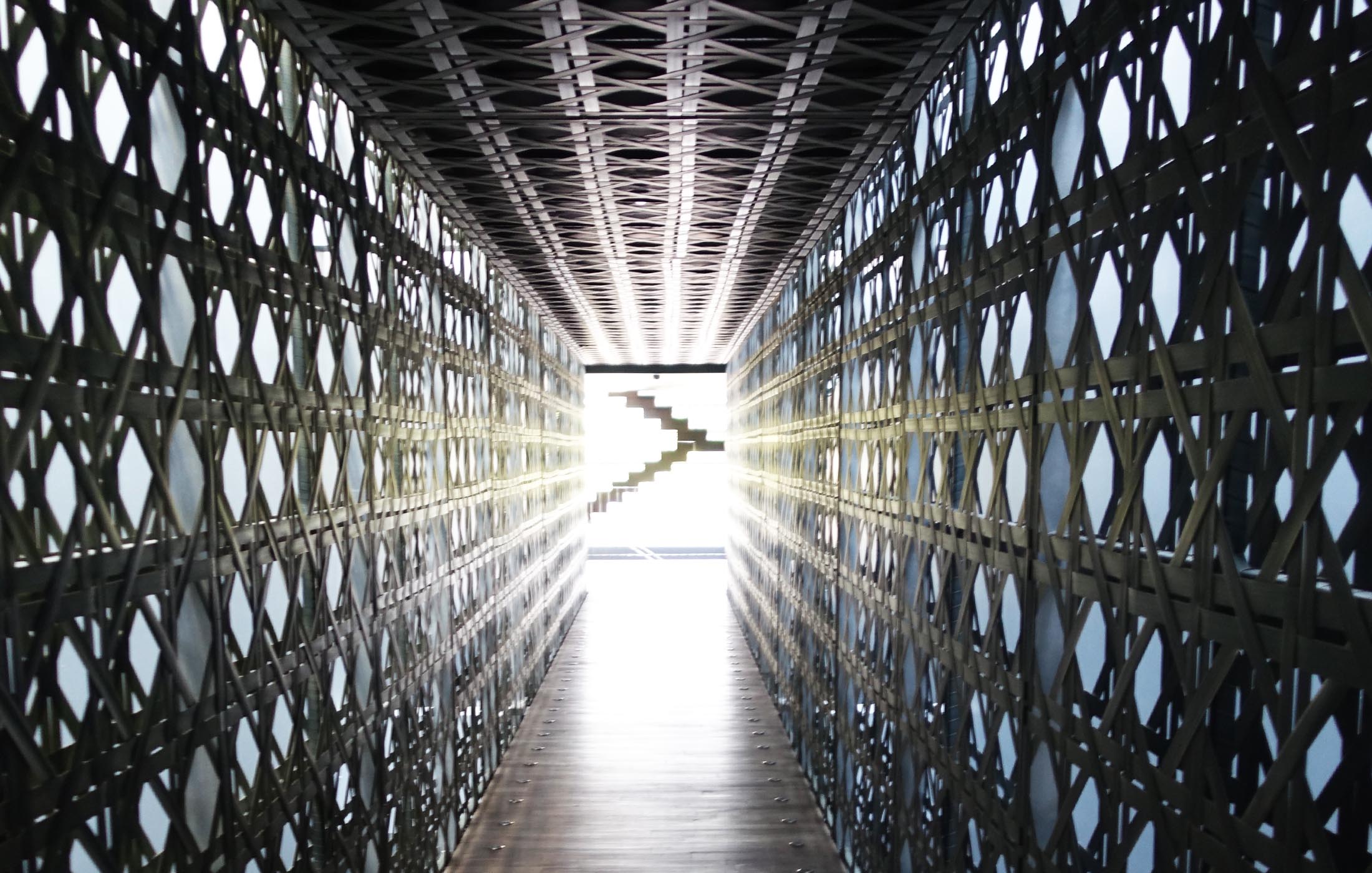 Langer, schmaler Flur mit strukturierter Metallgitterwand und Licht am Ende.