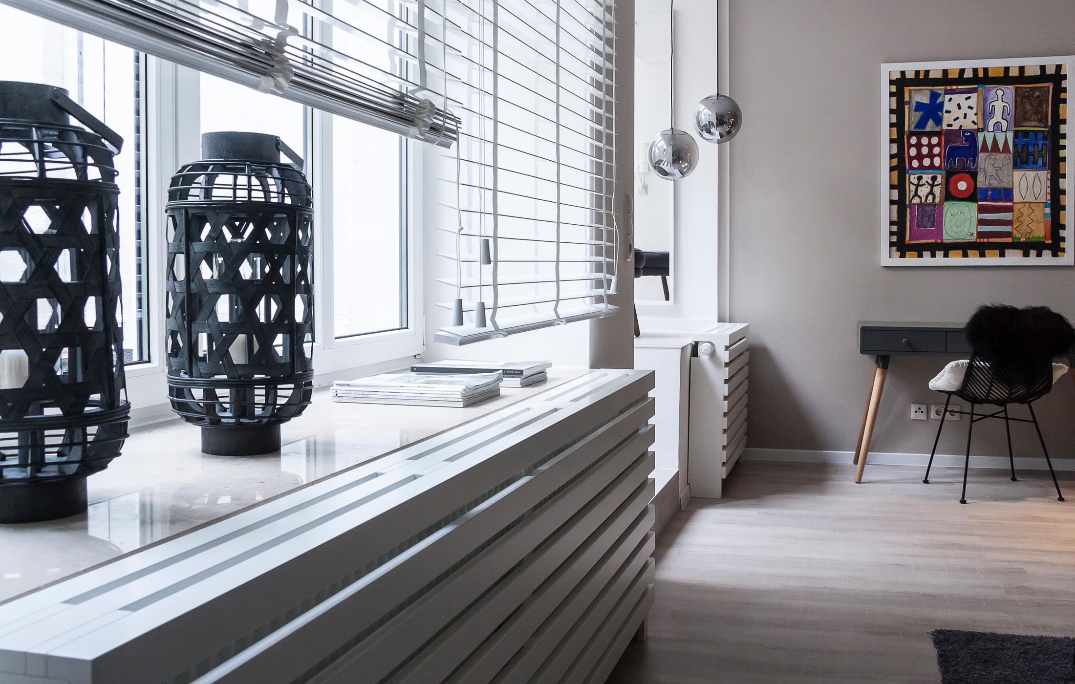 Ein freundlicher Wohnraum mit großen Fenstern, Holzdekor, schwarzer Deko und bunter Wandkunst.