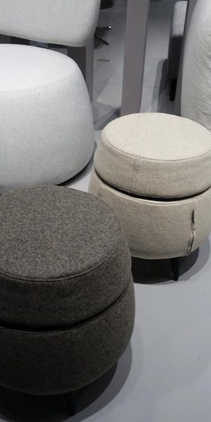 Drei runde Polsterhocker in Grau-, Beige- und Brauntönen auf hellem Boden neben einem Sofa.