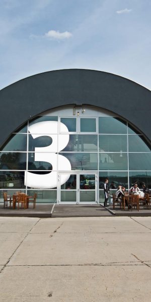 Frontansicht eines modernen Gebäudes mit großer Nummer drei an der Glasfassade und Holzsitzgruppen davor.