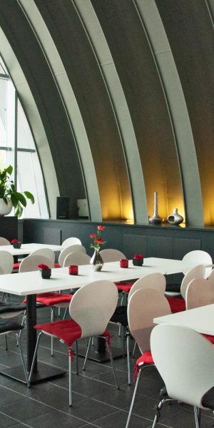 Blick von oben auf ein modernes, lichtdurchflutetes Bistro mit weißen Tischen, roten Sitzkissen und großen Pflanzen vor Panoramafenstern.