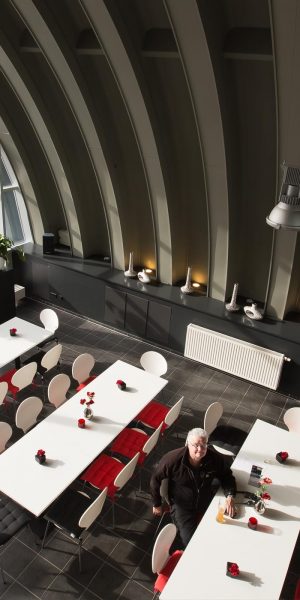 Blick von oben auf ein modernes, lichtdurchflutetes Bistro mit weißen Tischen, roten Sitzkissen und großen Pflanzen vor Panoramafenstern.