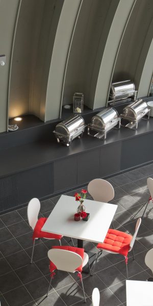Moderner Kantinenbereich mit weißen Tischen, roten Sitzkissen und großer Fensterfront.