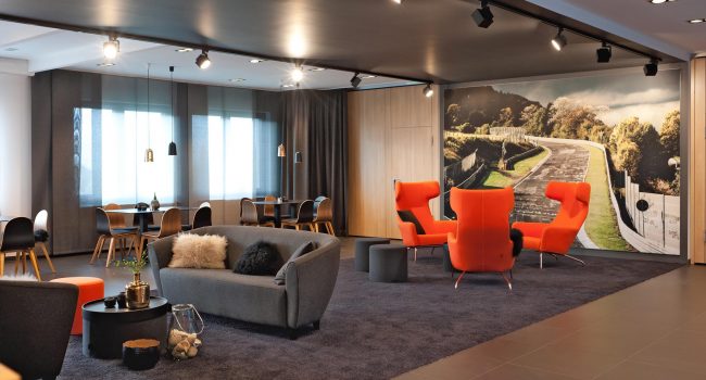 Loungebereich mit roten Sesseln und Sofa vor einer Fototapete der Nürburgring-Rennstrecke.