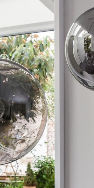 Nahaufnahme von zwei spiegelnden, kugelförmigen Pendelleuchten vor einem Fenster mit Gartenblick.