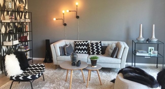 Ein stilvoll eingerichtetes Wohnzimmer mit grauer Couch, gemusterten Kissen, Designerlampen und einem modernen Schwarz-Weiß-Konzept.